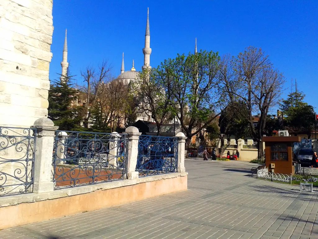 Örme Sütun'un dibi; beyaz taş babalar arasında kıvrımlı demir parmaklık, arkada Sultanahmet Camii'nin minareleri ve ağaçlar.