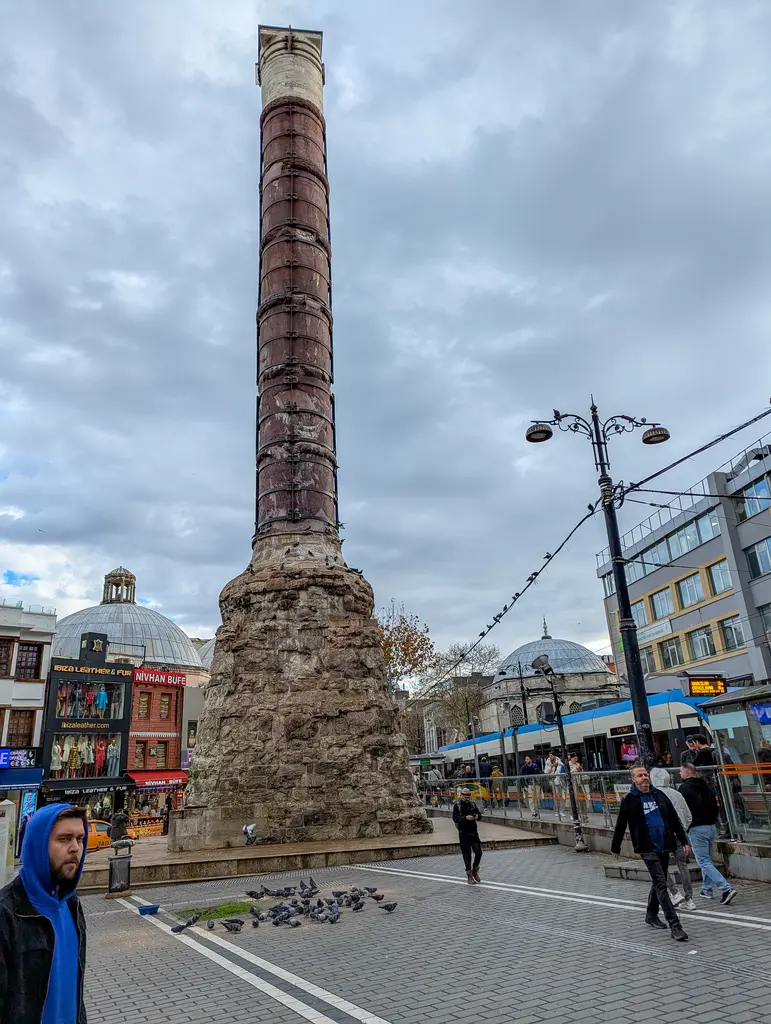 Çemberlitaş'ta kış günü sütun; soluk gökyüzü, taş kaide, önünden geçen yayalar ve arkadaki yapıların düz cepheleri.