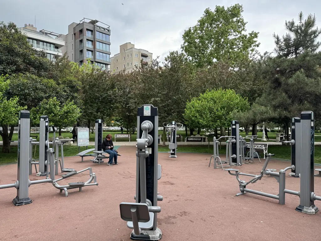 Göztepe 60. Yıl Parkı'nda açık hava spor aletleri; ağaçlar, çimenler, banklarda oturanlar ve arkada apartmanlar.