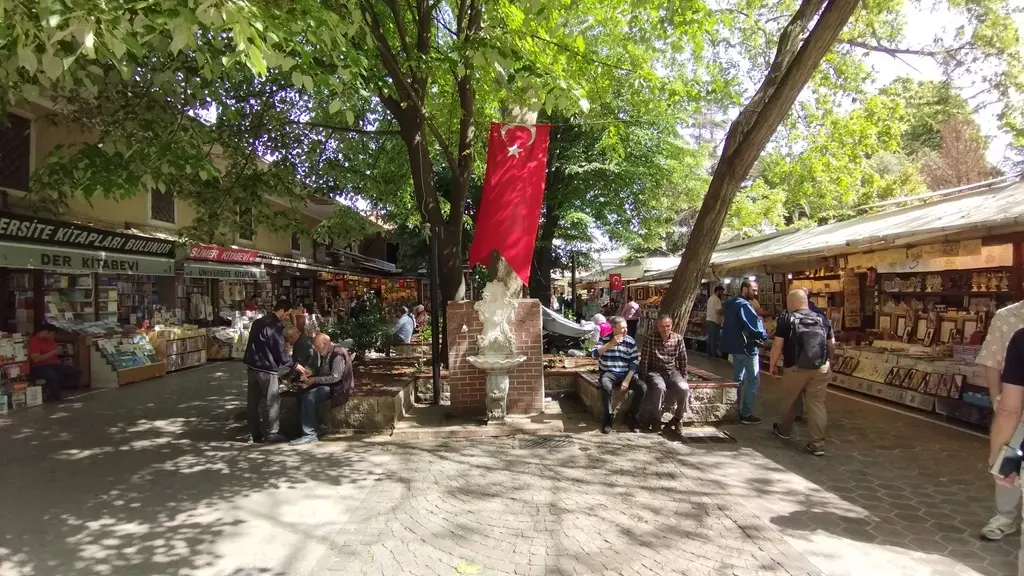 Sahaflar Çarşısı'nın göbeği; kırmızı tuğla ayağa oturtulmuş beyaz mermer çeşme, çınara asılı uzun Türk bayrağı, yanındaki taş sekide oturan iki kişi ve solda kitap seçen ziyaretçiler.