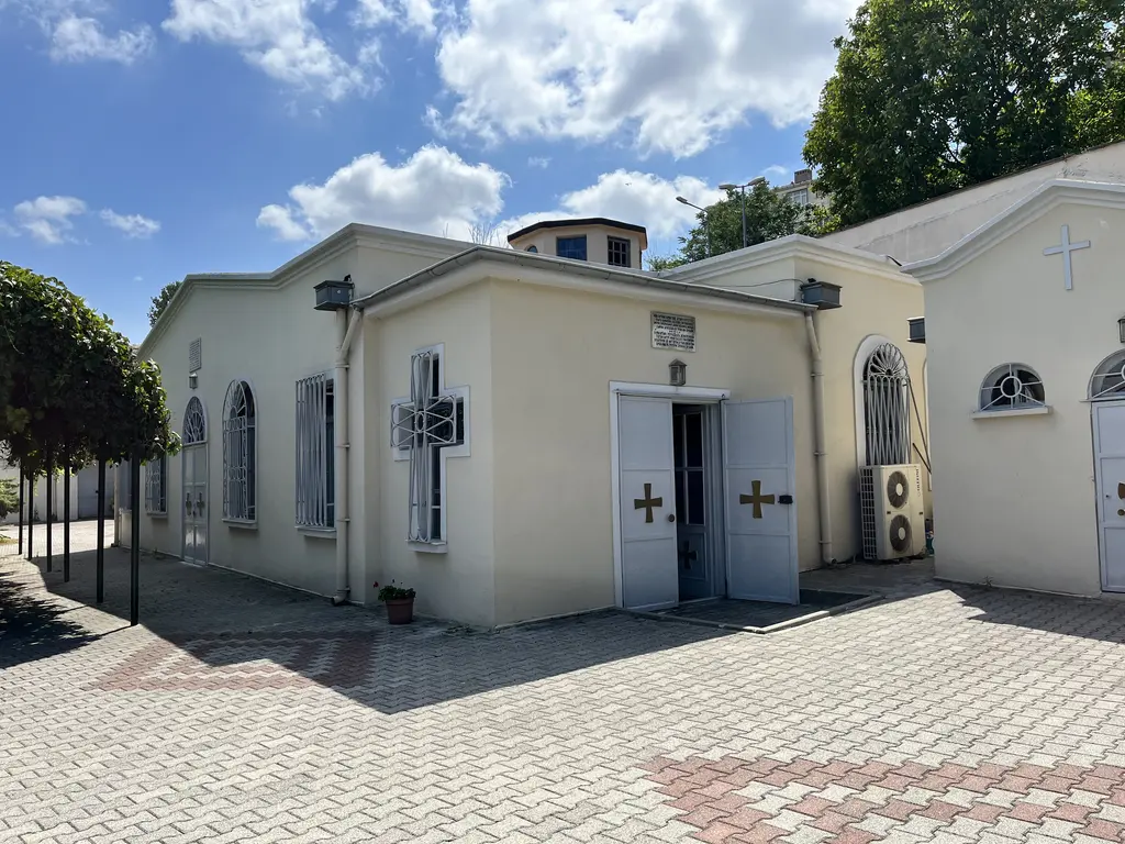 Güneşli bir günde avludan kilise; açık gri kapının iki kanadında altın haçlar, haç pencere, yanda klima dış üniteleri ve arkada ağaçlar.
