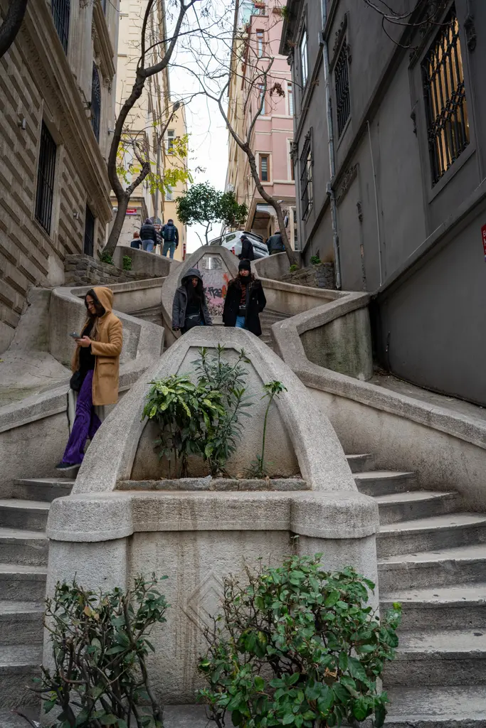 Kamondo Merdivenleri'nde yürüyen insanlar; kıvrımlı taş basamaklar, saksılardaki bitkiler ve yukarıda dar bir gökyüzü.