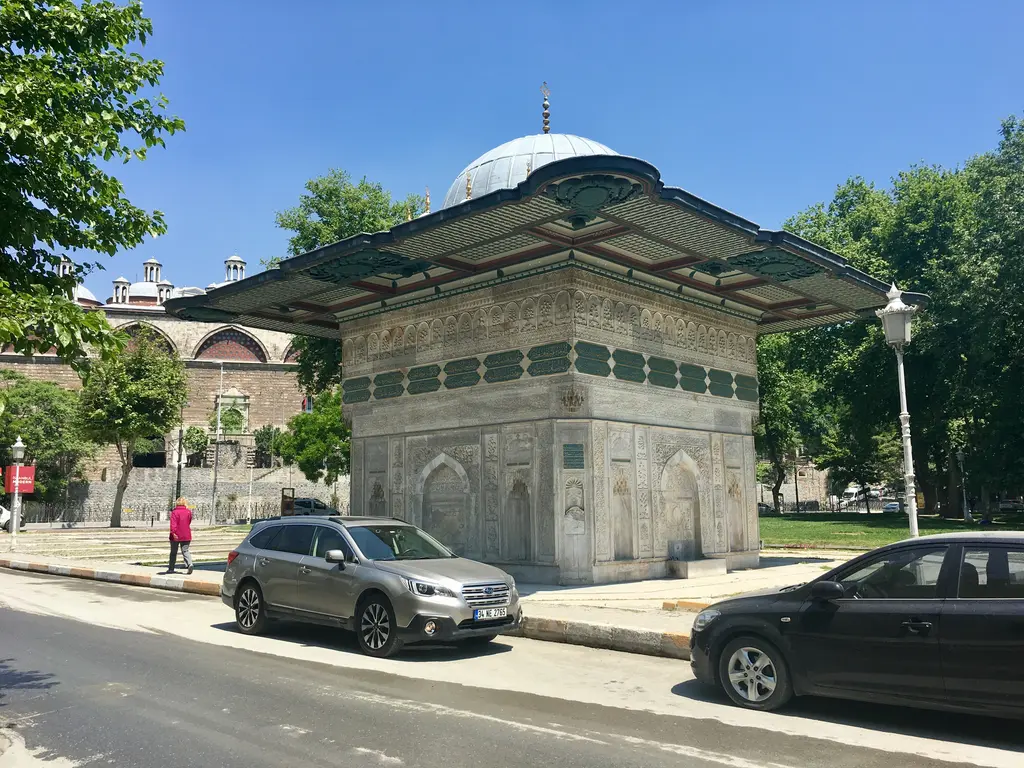 Tophane'de cadde kenarında çeşme; yeşil saçaklı mermer yapı, park etmiş arabalar ve arkada kubbeli bir yapı.