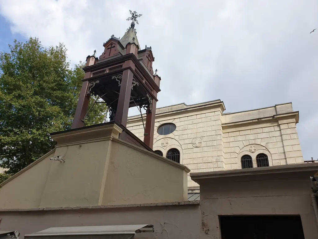 Surp Takavor Kilisesi'nin çan kulesi; ahşap kemerli kule, sivri külah, arkada kilisenin taş cephesi.