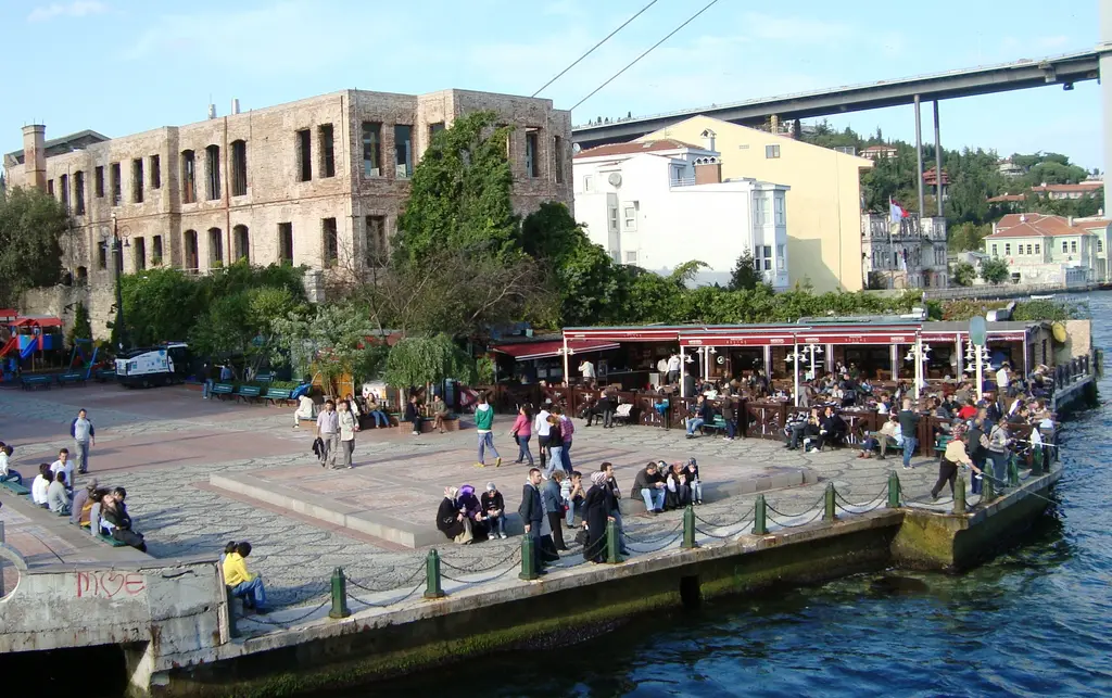 Boğaz'dan Ortaköy meydanı; kıyıda oturan insanlar, tenteli kafeler, yanık taş bina, ağaçlar ve üstte köprü.