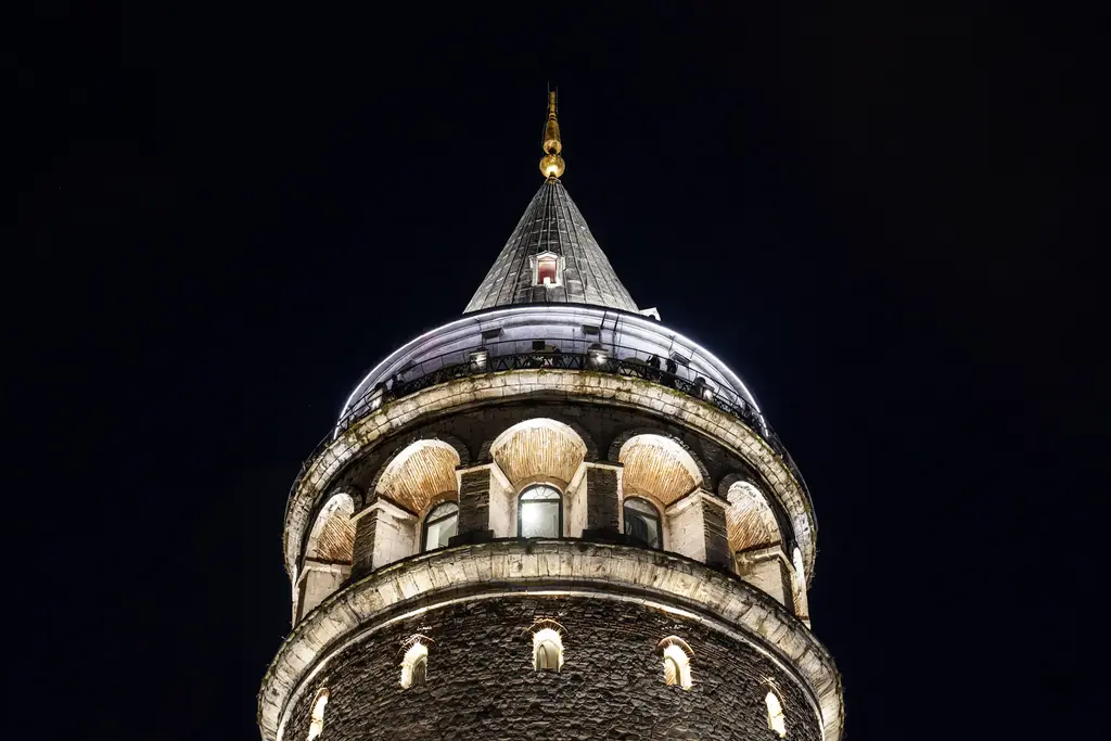 Gece aydınlatılmış Galata Kulesi'nin üst bölümü; ışıklı kemerli pencereler, sivri külah ile alem ve siyah gökyüzü.