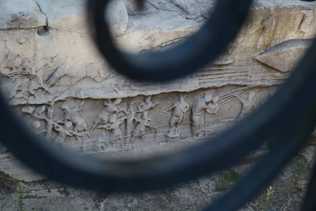 Dikilitaş kaidesinde kabartma; önde kıvrımlı siyah demir parmaklık, arkada koşan ve çeken figürlerin aşınmış izleri.