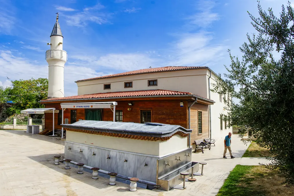 Kazlıçeşme Fatih Sultan Mehmet Camii; ahşap kaplamalı gövde, kiremit çatı, beyaz minare, önde üzeri kapalı şadırvan ve zeytin ağaçları.