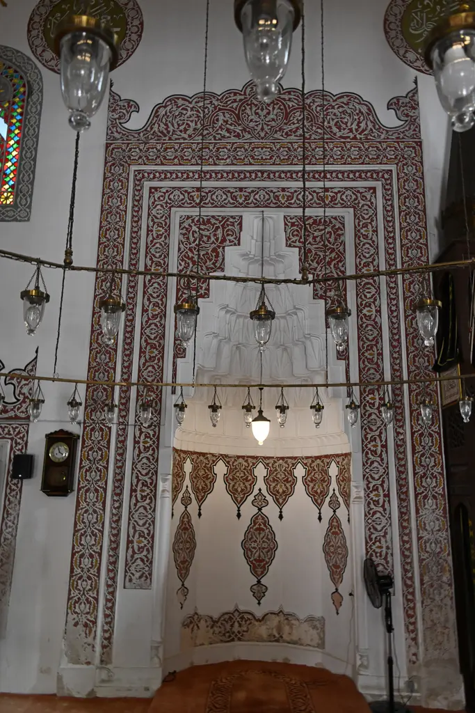 Firuz Ağa Camii'nin mihrabı; mukarnaslı alçı niş, çevresinde kırmızı kalem işi süslemeler, sarkan kandiller ve halı zemin.