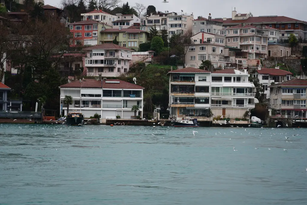 Çengelköy kıyısında suya inen beyaz ve krem renkli evler; yamaçta apartmanlar, ağaçlar ve önde dalgalı mavi su.