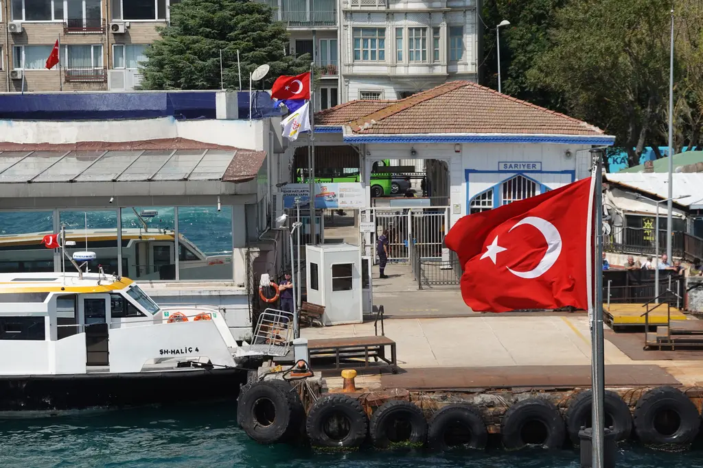 Sarıyer vapur iskelesi; mavi beyaz iskele binası, kiremit çatı, yanaşmış vapur ve önde Türk bayrağı.
