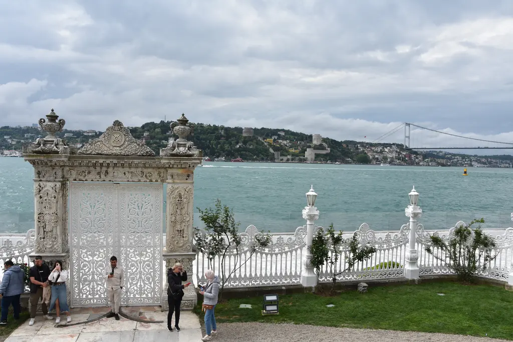 Küçüksu Kasrı'nın deniz kapısı; beyaz oymalı kapı, parmaklıklar, ziyaretçiler, arkada Boğaz, Rumeli Hisarı ve Fatih Sultan Mehmet Köprüsü.