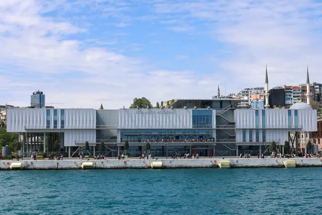 Boğaz'dan İstanbul Modern; kıyı boyunca uzanan açık gri, alçak ve uzun bina, önünde turkuaz deniz, arkada Beyoğlu yamacı.