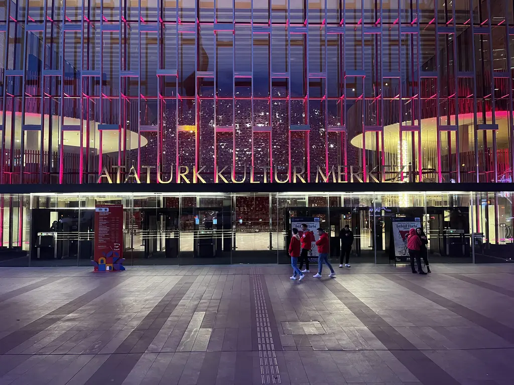 Atatürk Kültür Merkezi'nin girişi gece; ışıklı metal cephe, büyük harflerle bina adı, cam kapılar ve önde taş döşeli meydan.