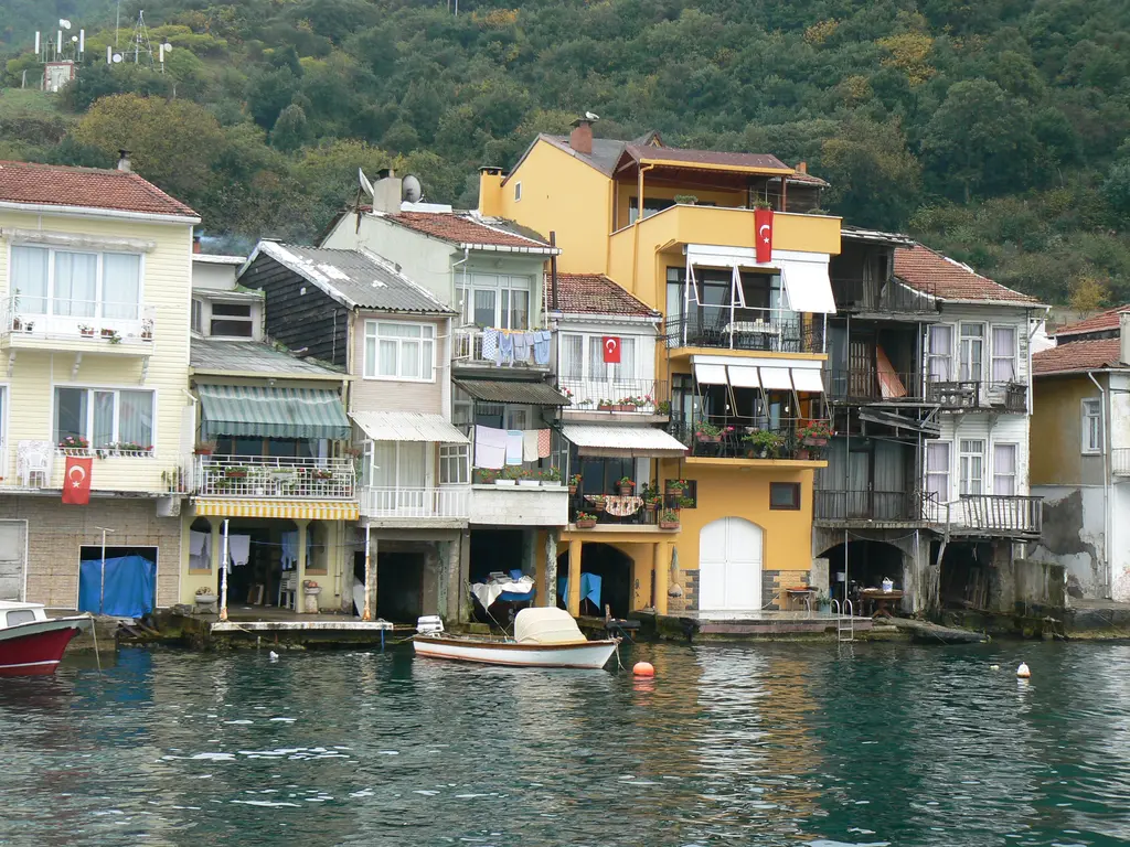 Anadolu Kavağı'nda suya inen renkli evler; sarı, beyaz ve krem cepheler, balkonlarda Türk bayrakları ve önde sandal.