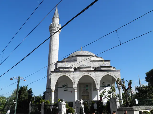 Sultanahmet'te Firuz Ağa Camii; beyaz taş cephe, üç kemerli son cemaat revakı, kurşun kubbe, ince minare ve önünden geçen tramvay telleri.