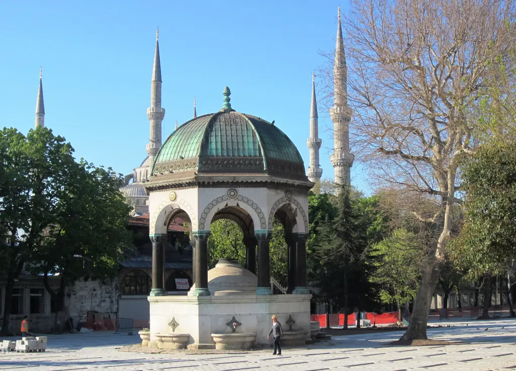 Alman Çeşmesi; yeşil kubbe, sütunlar ve kaide, arkada Sultanahmet Camii'nin minareleri, sağda yapraksız büyük bir çınar.