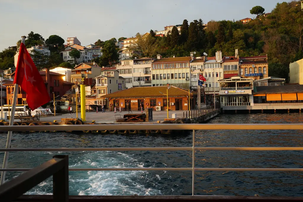 Beykoz iskelesi gün batımında; vapurun arka güvertesinden görünen iskele binası, bayrak ve kıyıdaki yapılar.