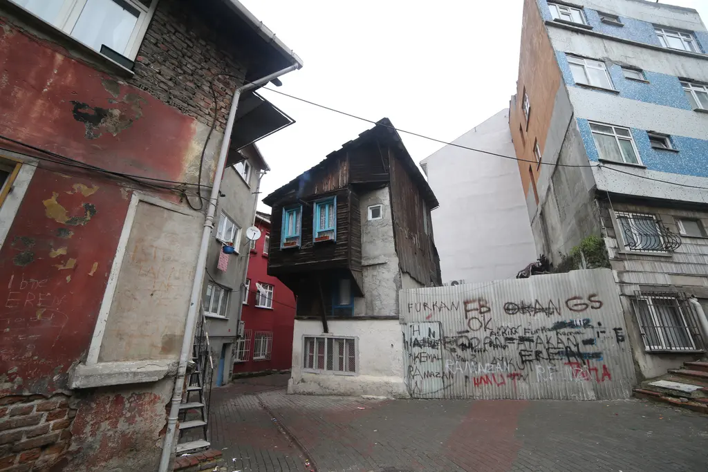 Fener'de dar sokak; solda kırmızı sıvalı ev, karşıda çıkmalı ahşap ev, grafitili teneke perde ve mavi apartman.