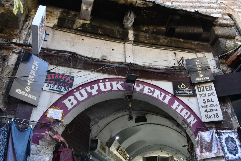 Büyük Yeni Han'ın sokaktan giriş kemeri; üstünde kırmızı Büyük Yeni Han tabelası, iki yanda şal ve eşarp satan dükkânlar.