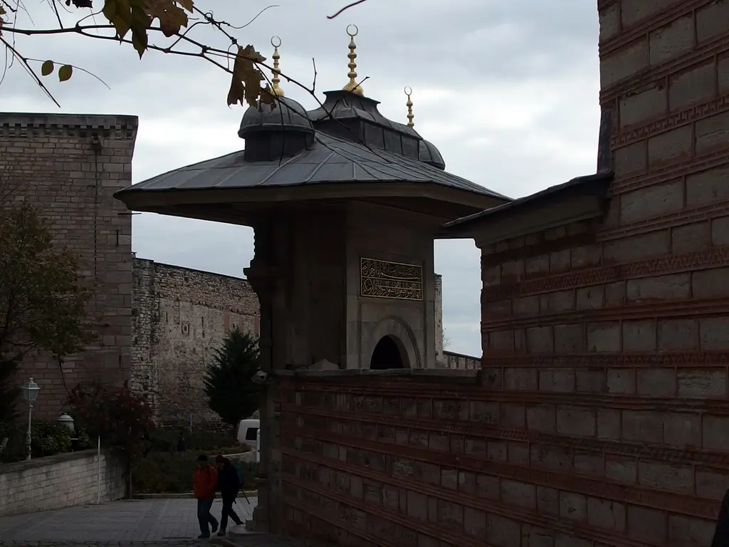Soğukçeşme Sokağı'nın üst ucunda iki duvar arasından görünen kurşun kubbeli çeşme çatısı, arkada sarayın yüksek suru ve bulutlu gök.
