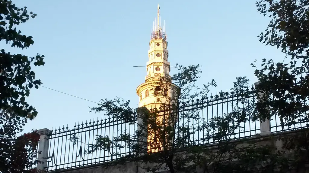 Gün batımında Beyazıt Kulesi; altın renginde parlayan taş gövde, önünde demir parmaklık ile ağaç dalları, arkada soluk gökyüzü.
