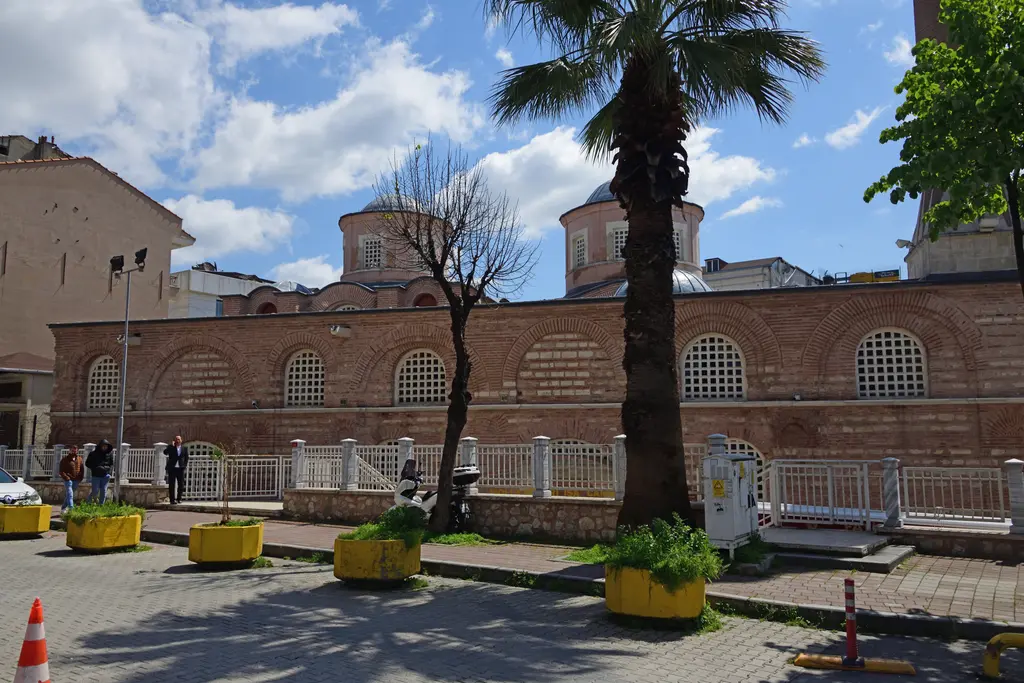 Fenari İsa Camii'nin batısındaki meydan; önde palmiye, yaprağını dökmüş ağaç ve sarı saksılar, arkada kemerli pencereli tuğla duvar ile iki kubbe birden.
