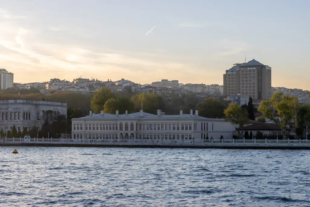 Denizden Dolmabahçe Sarayı; kıyı boyunca uzanan beyaz saray cephesi, arkasında ağaçlar ve tepedeki binalar, önde Boğaz'ın suyu.