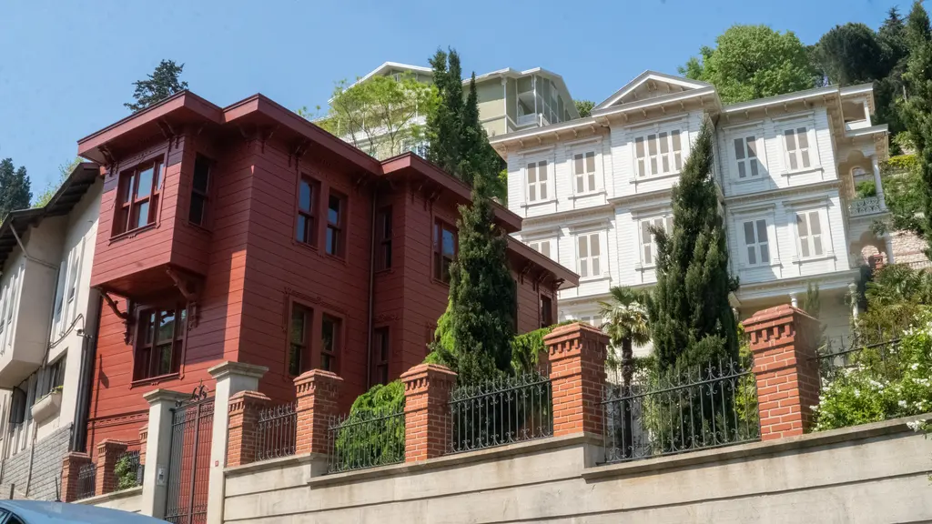 Bebek yakınında eski evler; kırmızı boyalı ahşap konak, yanında beyaz cepheli ev, servi ağaçları ve mavi gökyüzü.