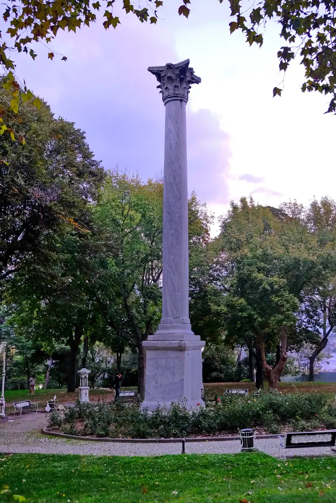 Gotlar Sütunu; mor tonlu bulutlu gök, kaidenin çevresinde çiçek tarhı, parke taş halka yol, banklar ve yeşil ağaçlar.