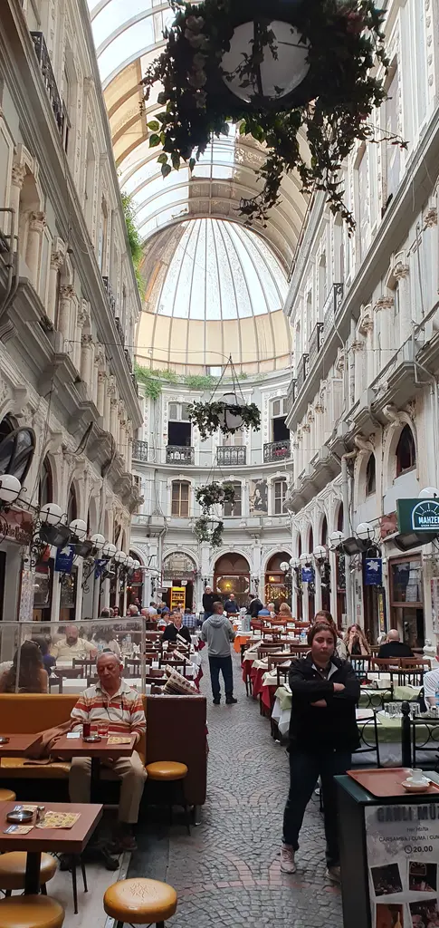 Çiçek Pasajı'nın içi; tavandan sarkan çiçek sepeti, cam çatının altında masalar, iki yanda süslü cepheler ve uzakta rotonda.