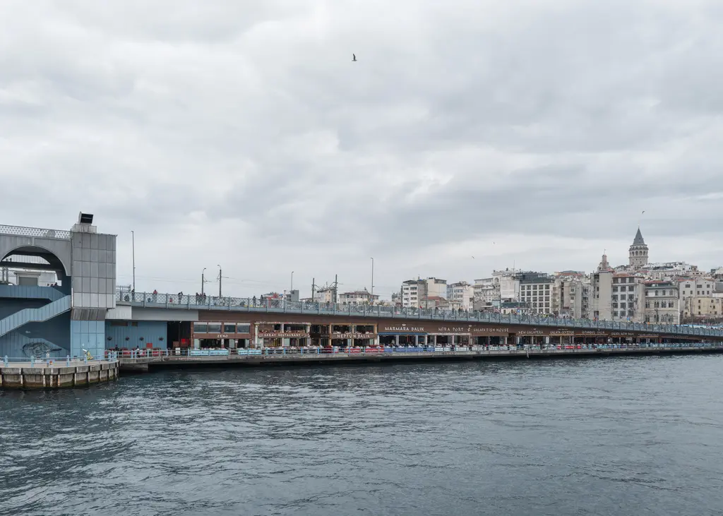 Haliç'in ağzında Galata Köprüsü; kapalı gökyüzü, gri su, köprünün uzun gövdesi ve arkada Karaköy'ün binaları.