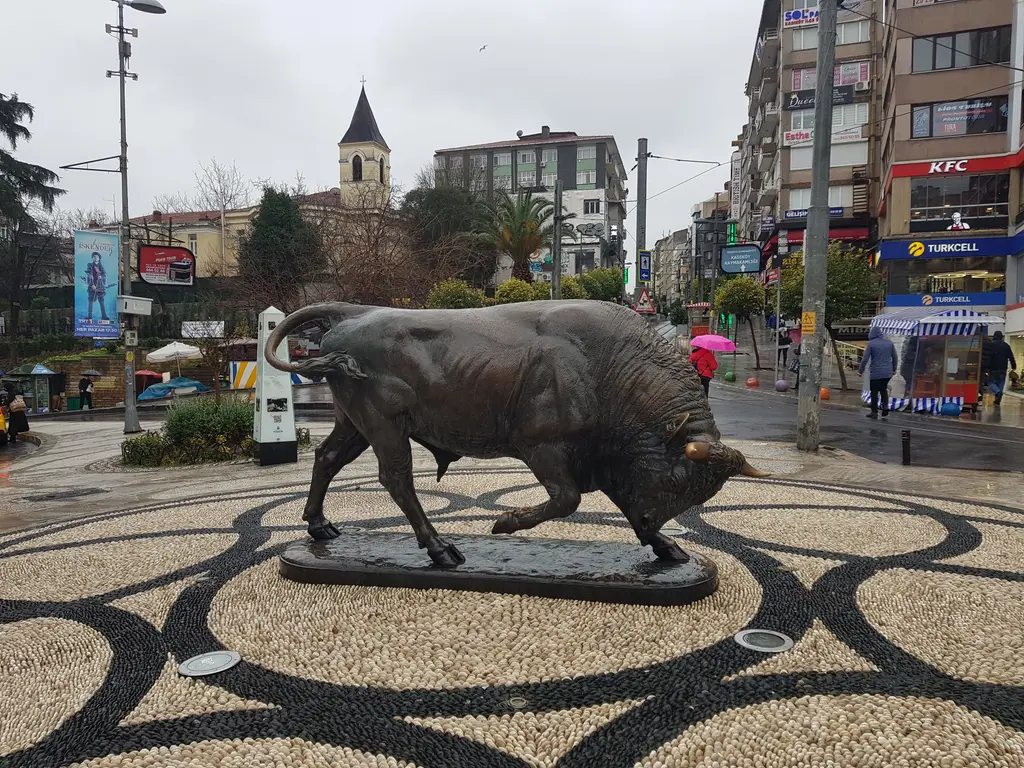 Kadıköy'de boğa heykeli; mozaik taş döşeli meydanda bronz boğa, arkada dükkânlar ve yürüyen insanlar.