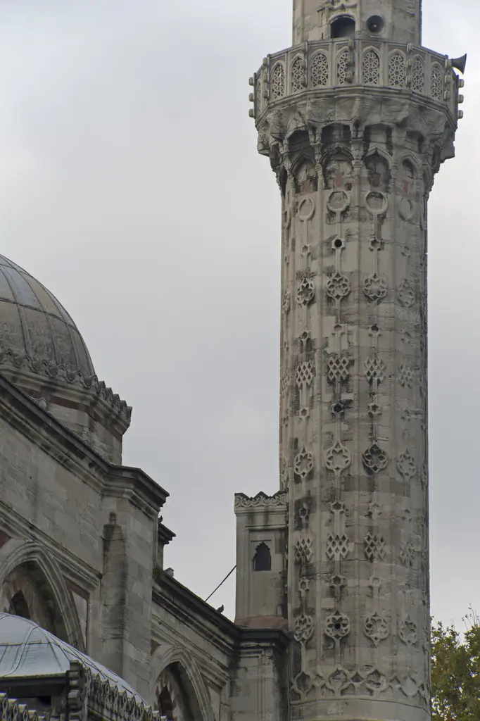 Şehzade Camii'nin minaresi yakın planda; gövdeyi baştan aşağı kaplayan kabartma geometrik geçmeler ve rozetler, şerefe altında mukarnas dizisi, gri gökyüzü.