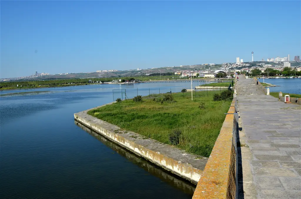 Kanuni Sultan Süleyman Köprüsü'nün yanında kanal; taş rıhtımla çevrili yeşil dil, mavi su ve köprü sırtının kenarı.