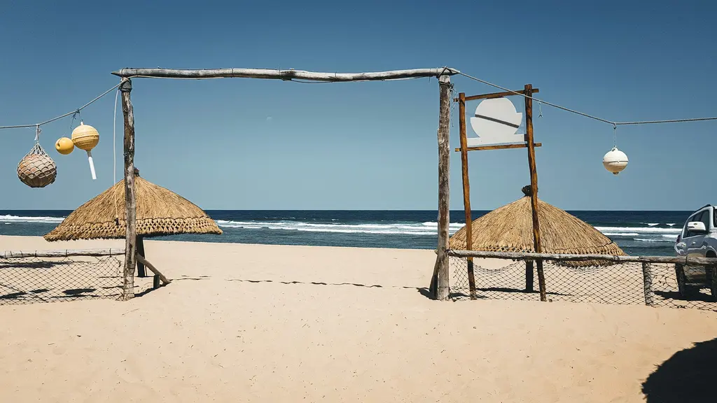 Mozambik'te bir plaj girişi; ahşap direklerden bir geçit, hasır şemsiyeler ve şamandıralar, arkada okyanus dalgaları ve bir arazi aracı.