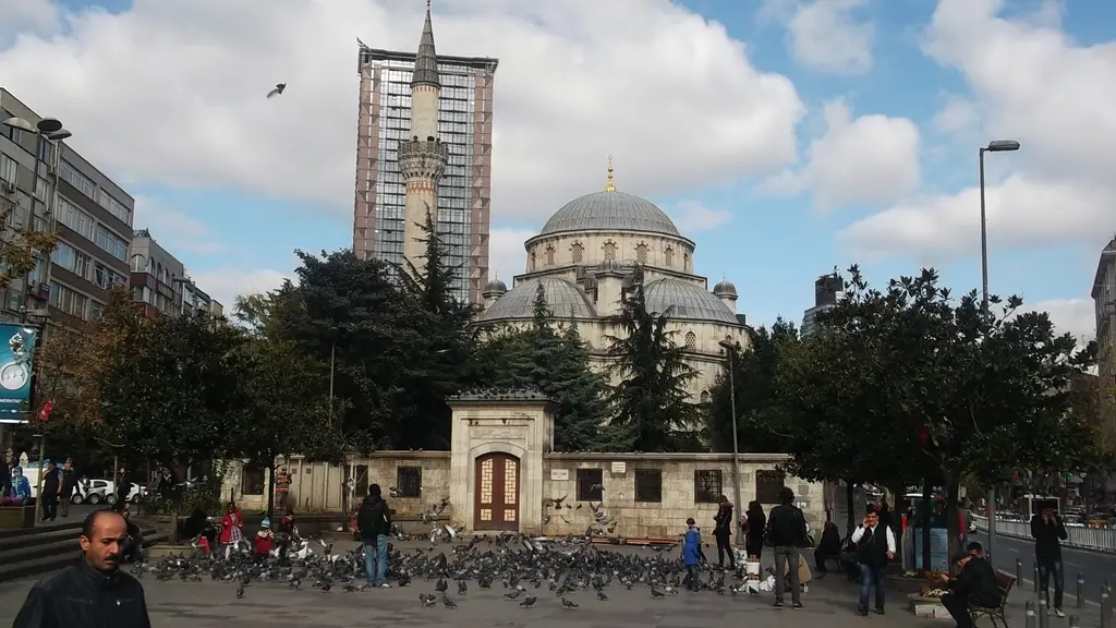 Şişli Camii'nin avlu duvarı ve önündeki meydan; güvercinler, yürüyen insanlar, ağaçlar ve arkada kubbeler ile minare.