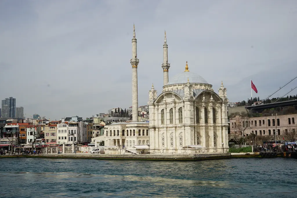 Denizden Ortaköy Camii ve semt; beyaz cami, minareler, arkada yamaca yayılan Ortaköy evleri ve önde iskele.
