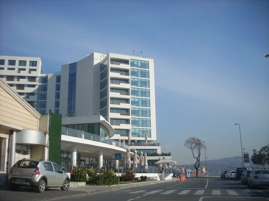 Tarabya'da kıyı yolu; park etmiş araçlar, kaldırımda ince bir ağaç ve arkada Büyük Tarabya Oteli'nin blokları.