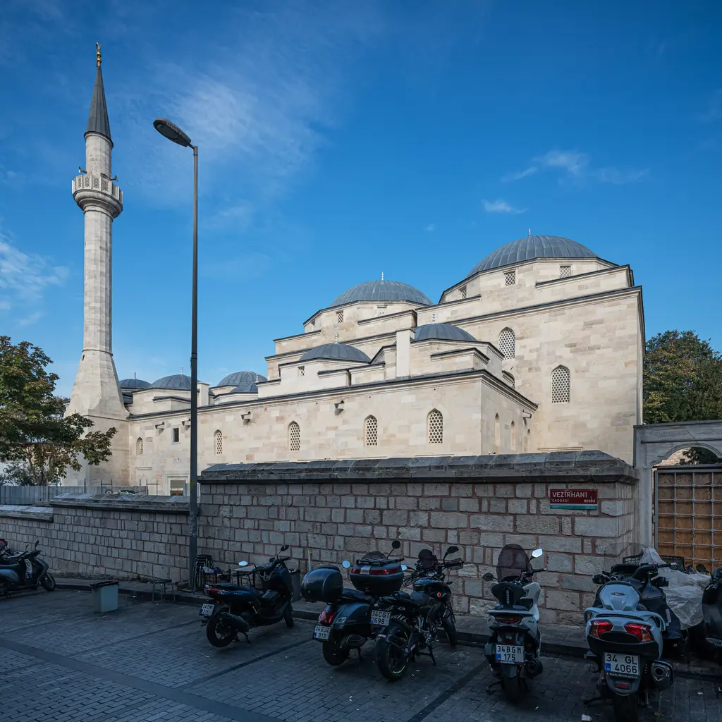 Mahmut Paşa Camii; solda ince taş minare ve sokak lambası, taş avlu duvarı, arkada kubbeler ve önde park edilmiş motosikletler.