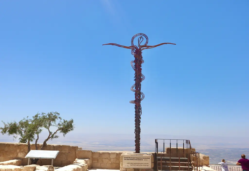 Nebo Dağı'nın tepesinde metalden yapılmış yılanlı haç anıtı; anıtın çevresindeki taş teras, korkuluklar, yanındaki tek ağaç ve aşağıda uzanan geniş vadi manzarası.