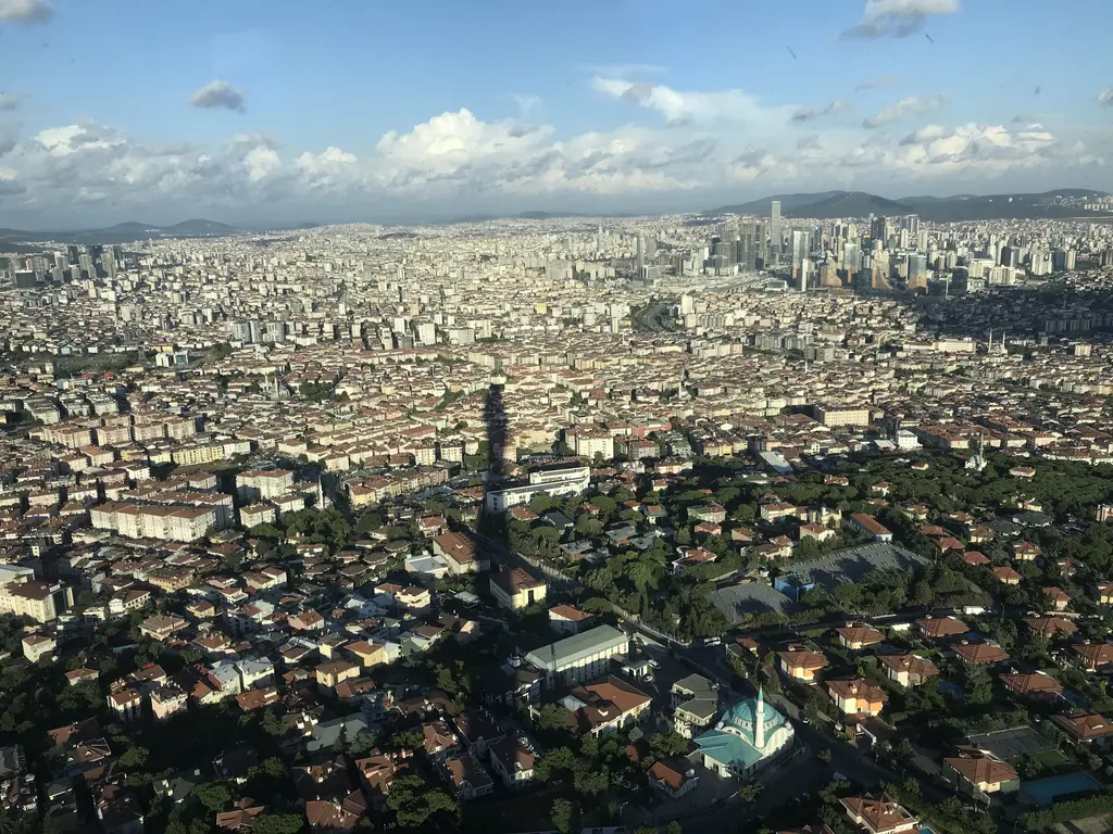 Çamlıca Kulesi'nden kuşbakışı İstanbul; sıkışık yerleşim, uzanan şehir, ufukta deniz ve aşağıda kulenin gölgesi.