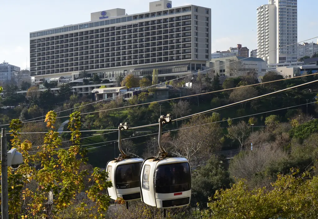 Maçka teleferiğinin eski tip iki beyaz kabini ağaçlı vadinin üstünde; arkada Hilton oteli, bir gökdelen ve açık gökyüzü.