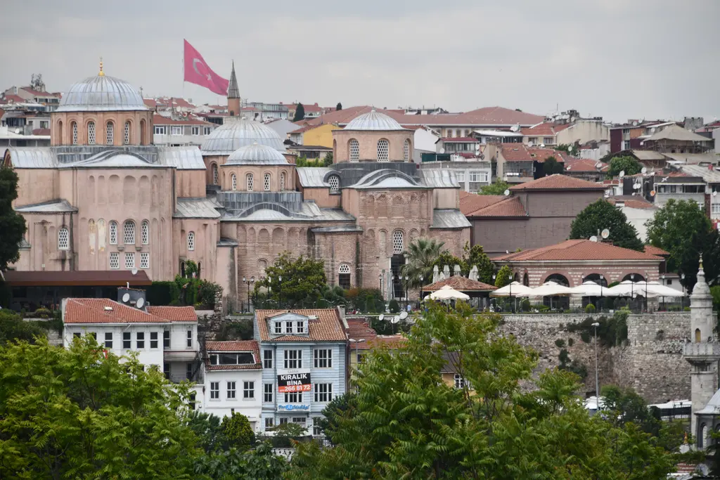 Zeyrek yamacına bakış; kurşun kubbeli, taş ve tuğla örgülü Zeyrek Camii, önde kırmızı kiremitli evler, ağaçlar ve Türk bayrağı.