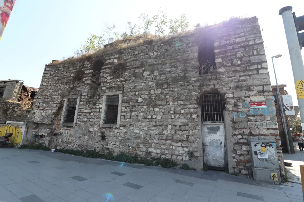 Ayakapı Hamamı'nın taş cephesi; demir parmaklıklı pencereler, çatıda bitkiler, kaldırım ve duvarda afişler.