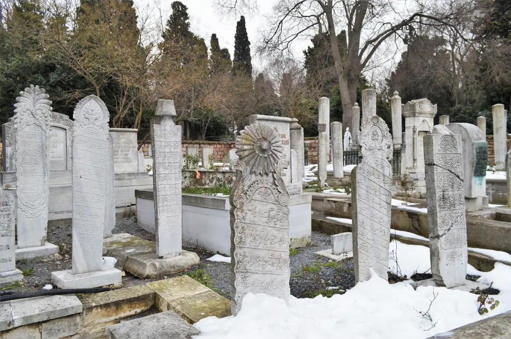 Karla kaplı mezarlıkta eğimli duran eski mezar taşları; kavuklu ve çiçek kabartmalı başlıklar, arkada selviler.