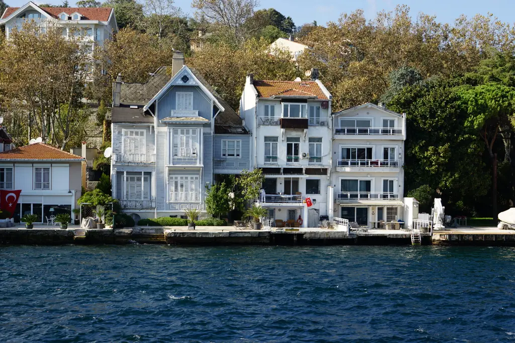 Saip Özden Yalısı; beyaz ve açık mavi ahşap cepheler, sivri çatılar, Türk bayrağı ve önde rıhtım.