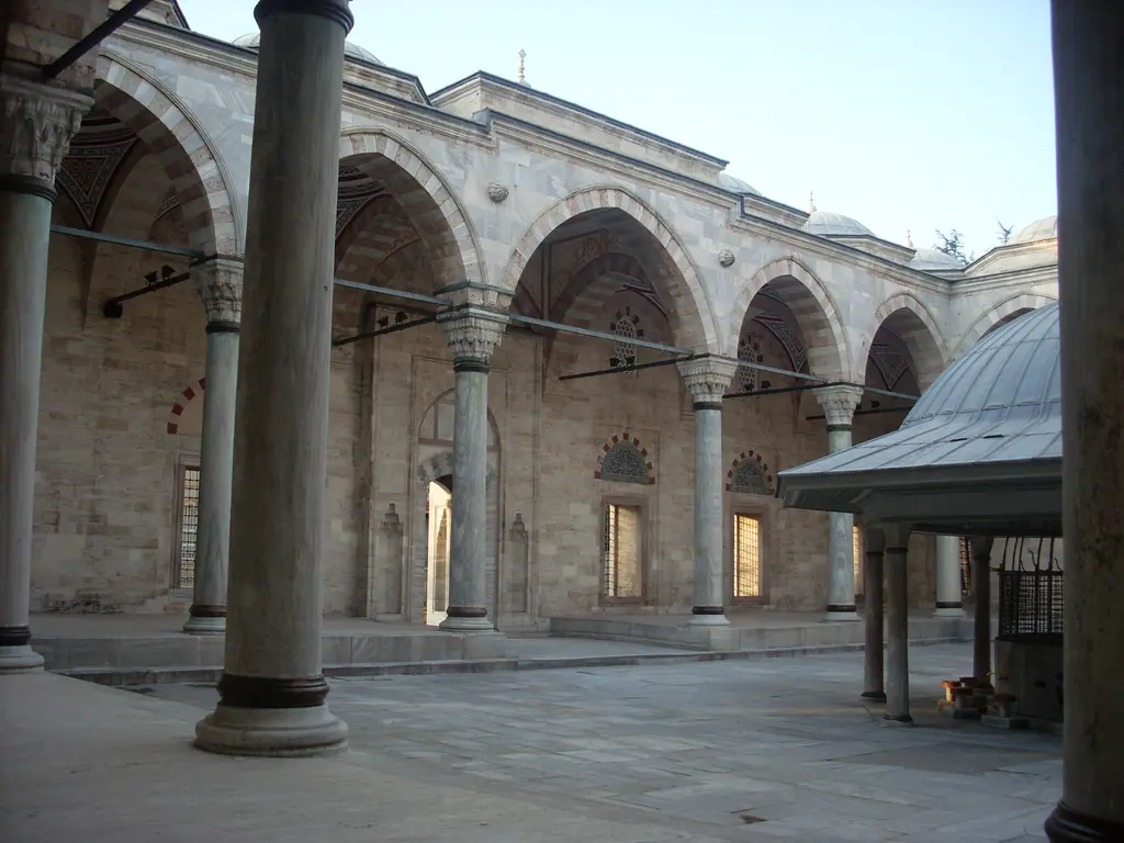 Yavuz Sultan Selim Camii'nde revağın içinden bakış; kalın mermer sütunlar, sivri kemer dizisi, sağda şadırvanın saçağı ve ıslak mermer zemin.
