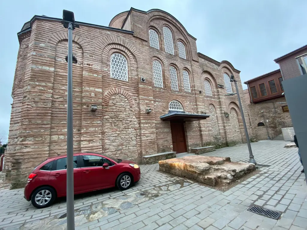 Zeyrek Camii sokaktan; iki katlı kemerli pencere dizisi, tuğla ile taşın almaşık örüldüğü duvar, kaldırımda kırmızı otomobil ve parke taşlı eğimli yol.