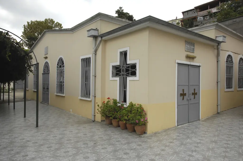 Kilisenin sarı ve krem boyalı yan cephesi; kemerli demir parmaklıklı pencereler, haç biçimli pencere, altın haçlı gri kapı ve saksılarda sardunyalar.